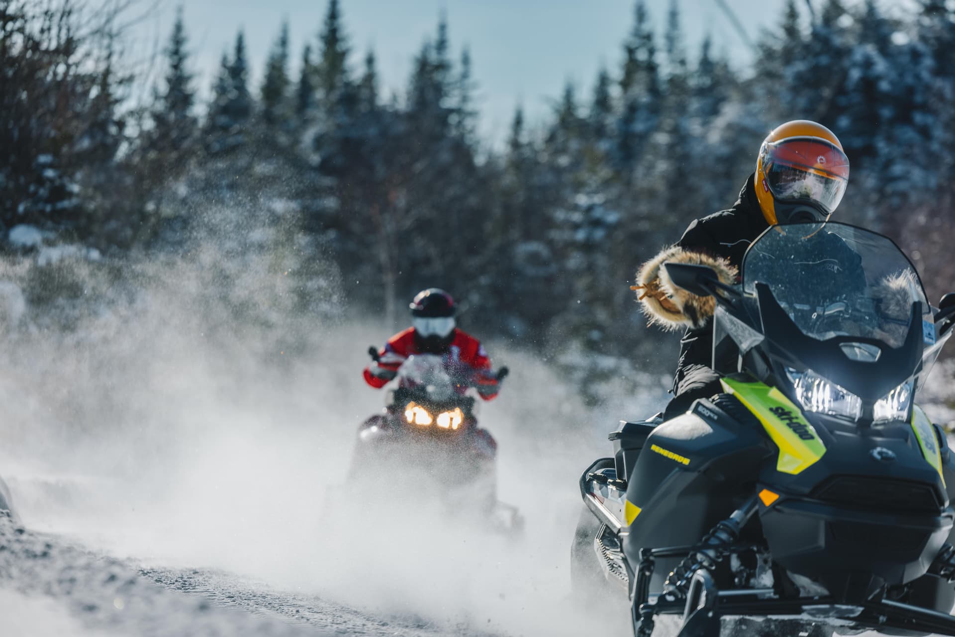 Close-up of two snowmobilers riding on a snow-covered trail, with snow-covered trees in the background.