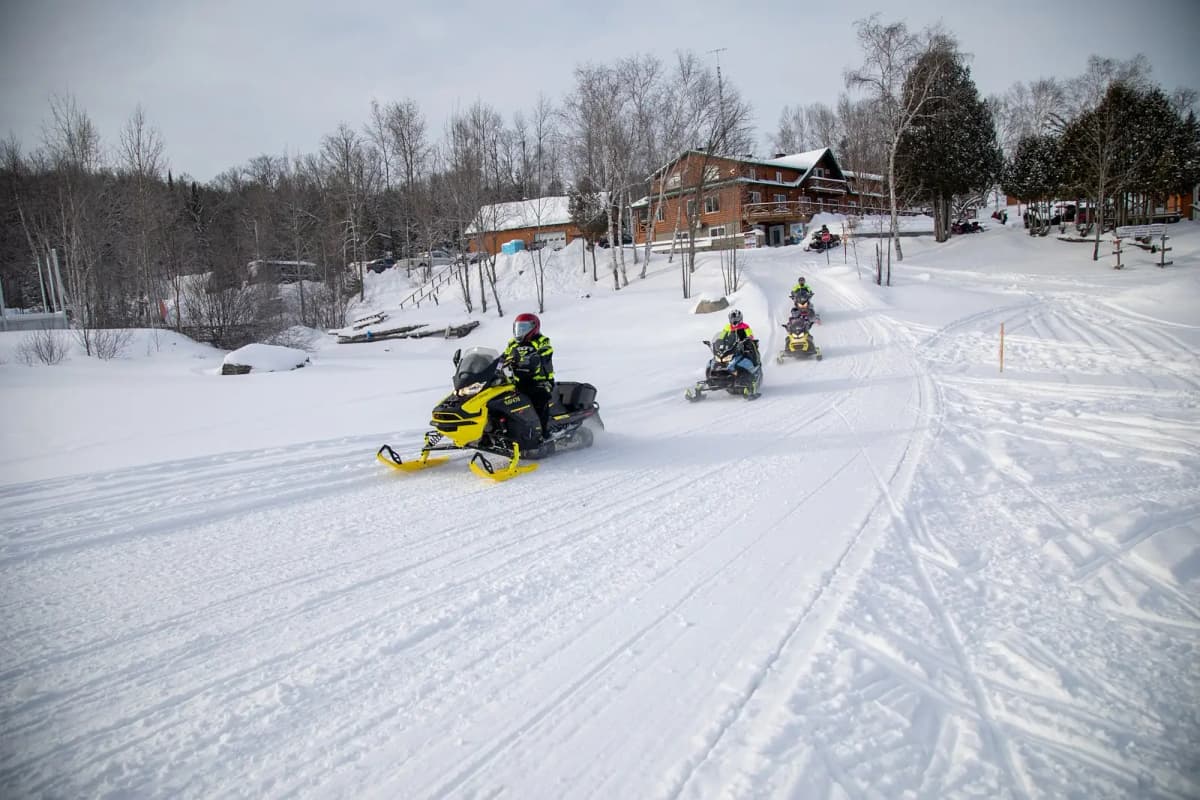 Un groupe de motoneigistes roule sur un sentier enneigé près d’un chalet en bois, dans les Laurentides, une région reconnue comme l’une des meilleures destinations pour la motoneige.