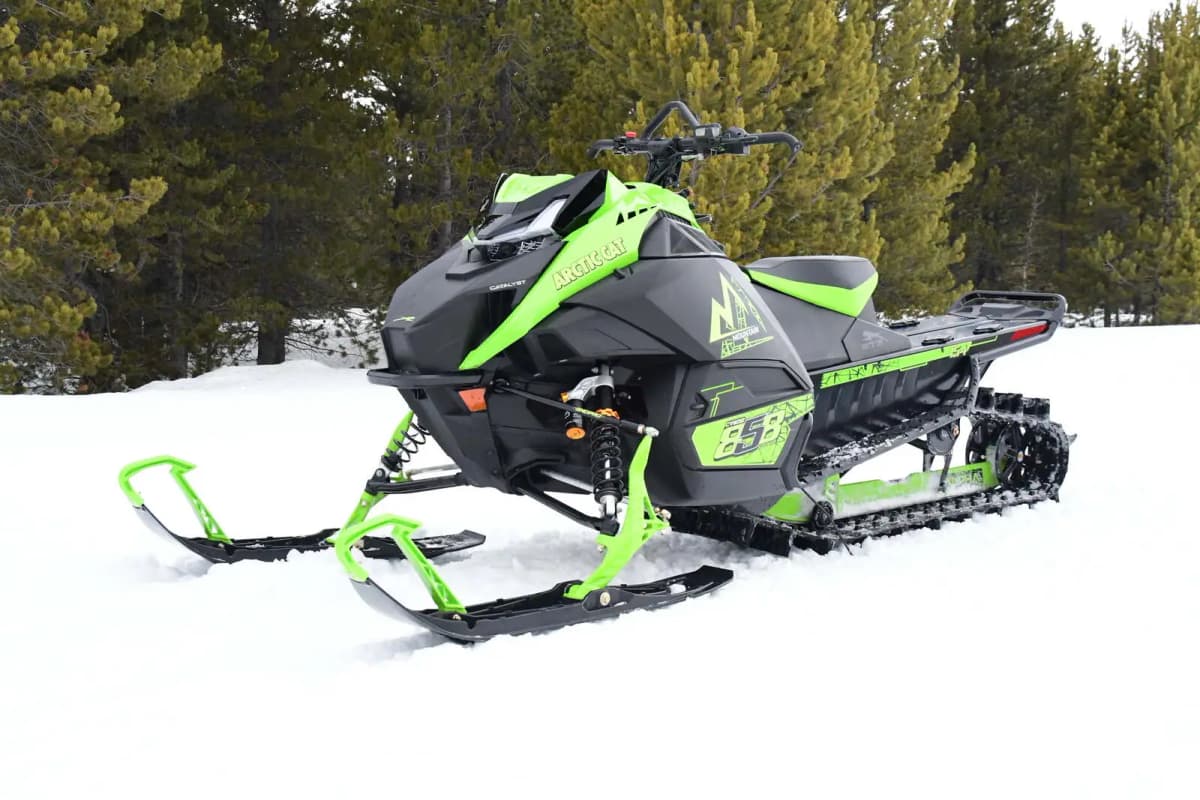 Gros plan d’une M 858 Alpha One Sno Pro verte, stationnée sur un terrain enneigé avec une forêt de sapins en arrière-plan.