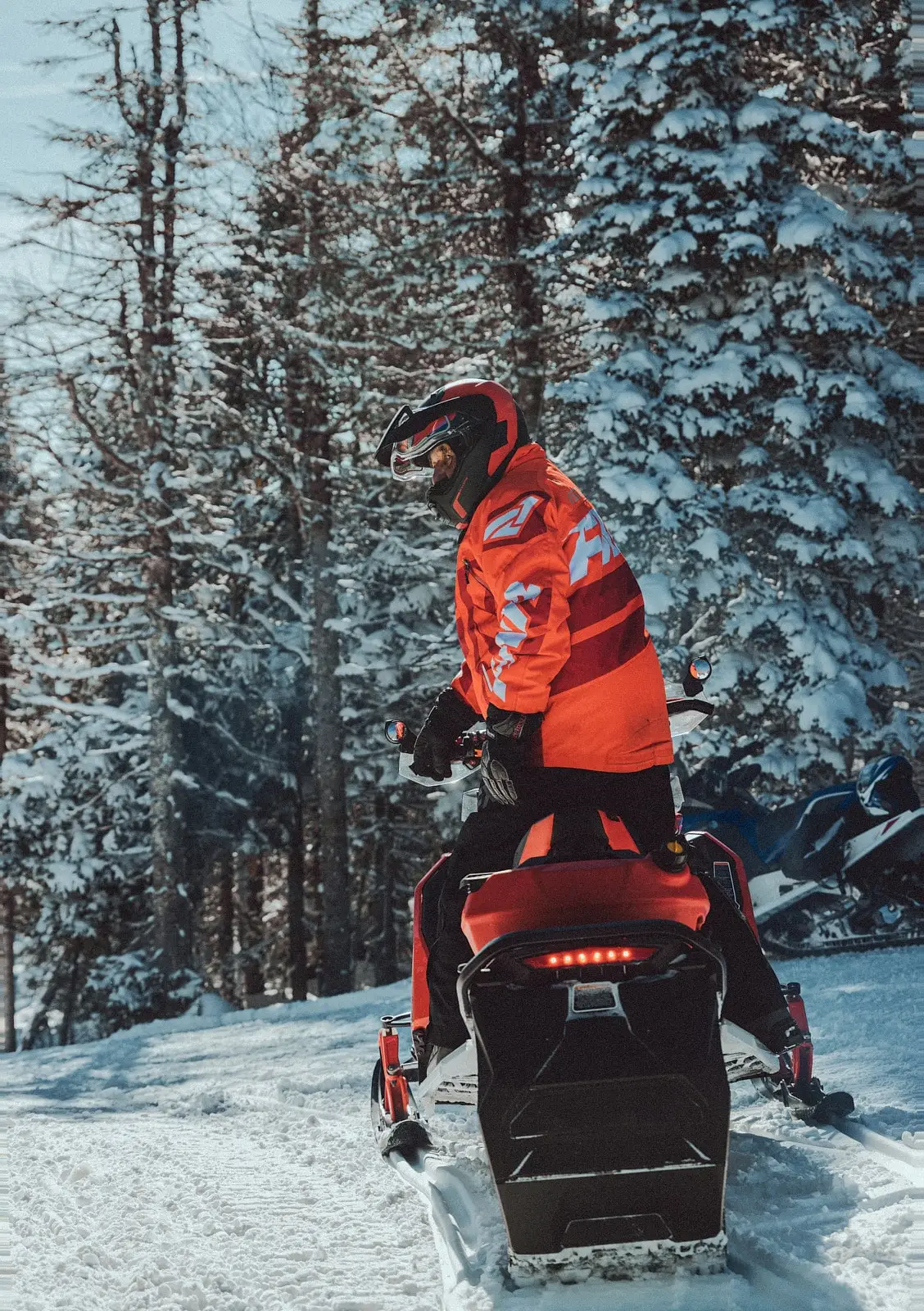 Un motoneigiste en tenue rouge se tient debout sur sa motoneige, prêt à parcourir un sentier enneigé à travers une forêt.