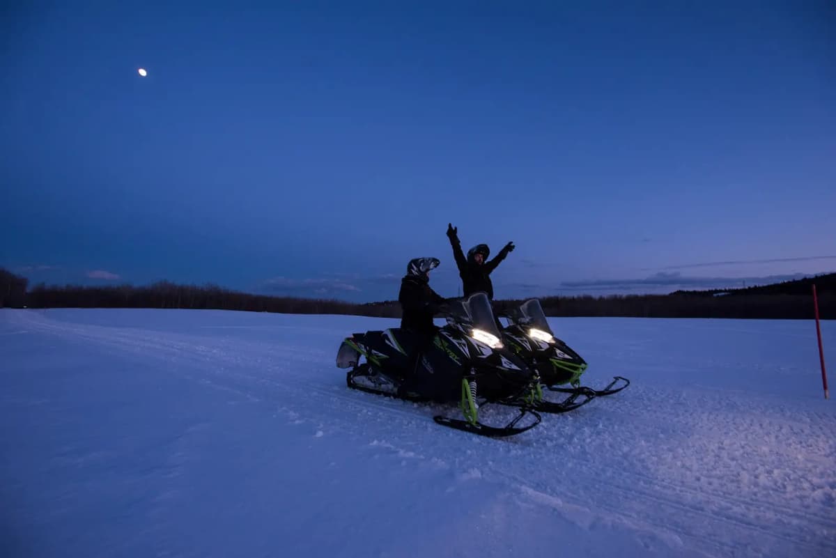 Deux motoneigistes en pleine randonnée sur les sentiers de motoneige en Abitibi-Témiscamingue, s’arrêtent sous un ciel nocturne, l’un levant les bras en signe de joie.