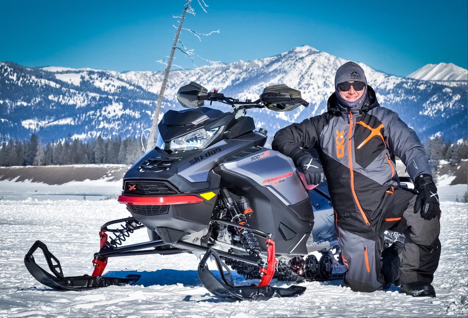 a man wearing the ckx elevation mono-suit kneeling by his snowmobile