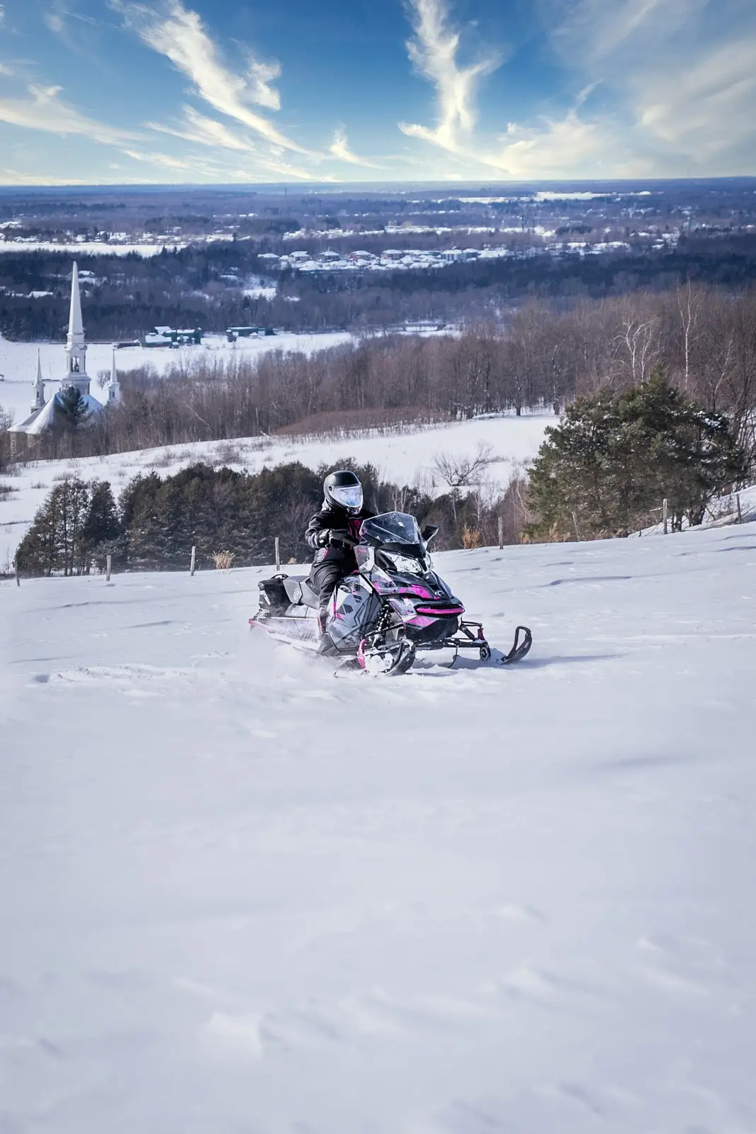 A snowmobile rider walks through a snow-covered field with a panoramic view of a church and the winter landscapes of Centre-du-Québec in the background.