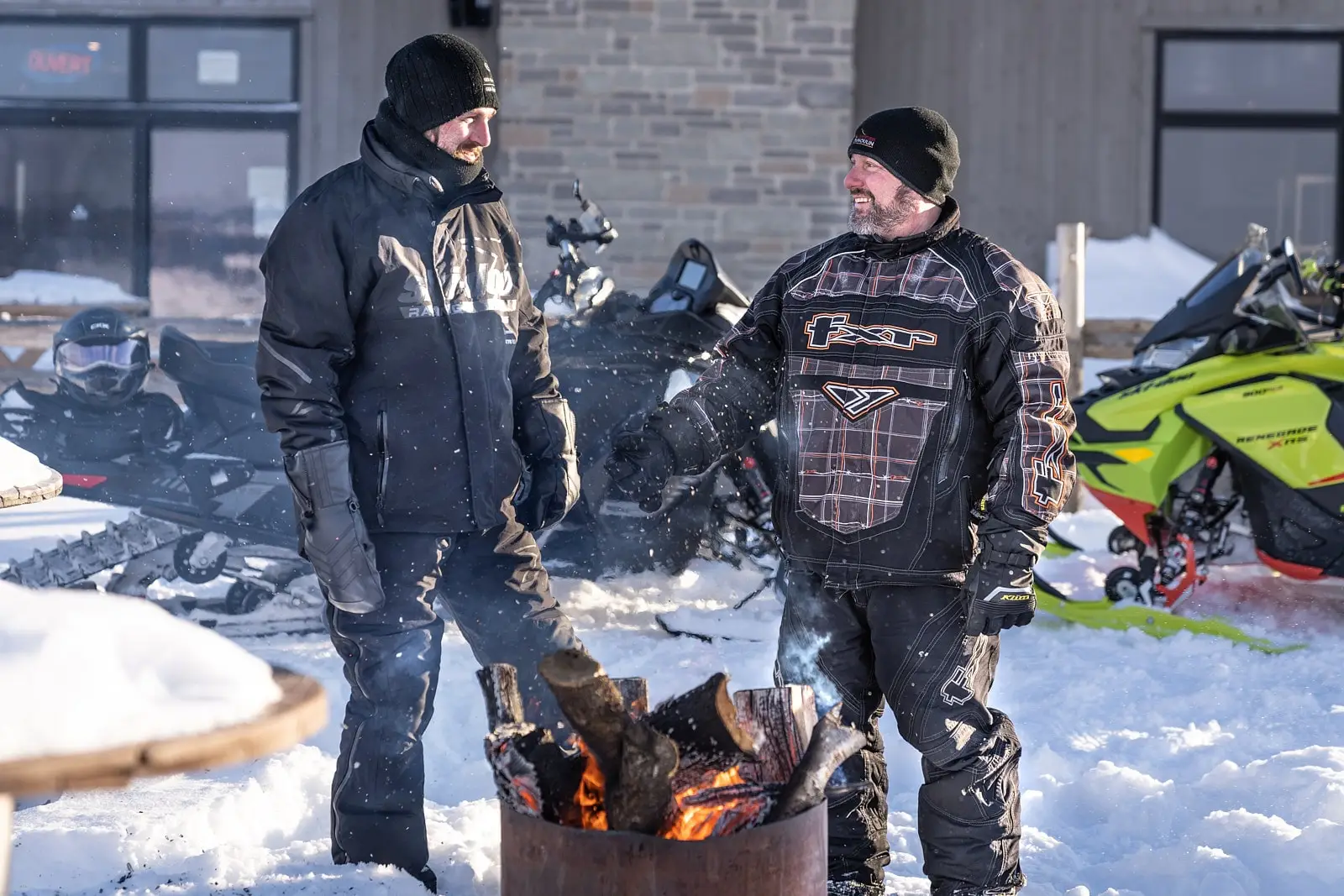 Two men in snowmobile uniforms are talking outside at a campfire, with snowmobiles parked in the background on the snow.