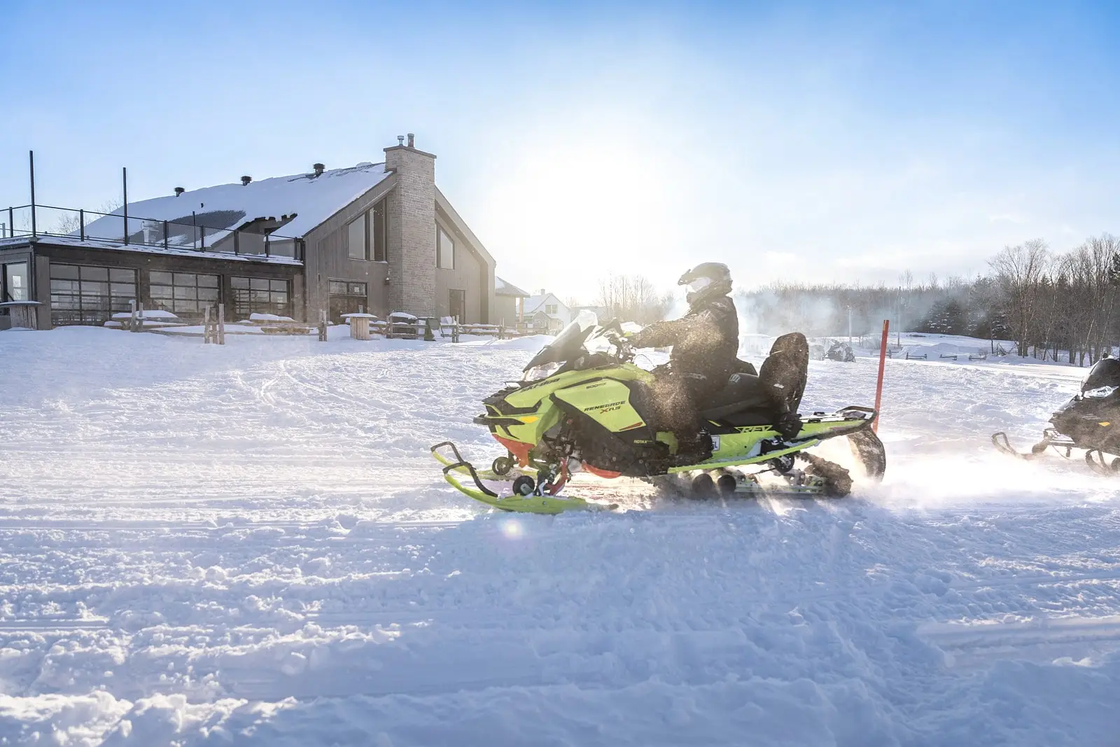 A snowmobile rider on a yellow fluo snowmobile passes in front of the large snow-covered Grange Pardue chalet, illuminated by the sun.