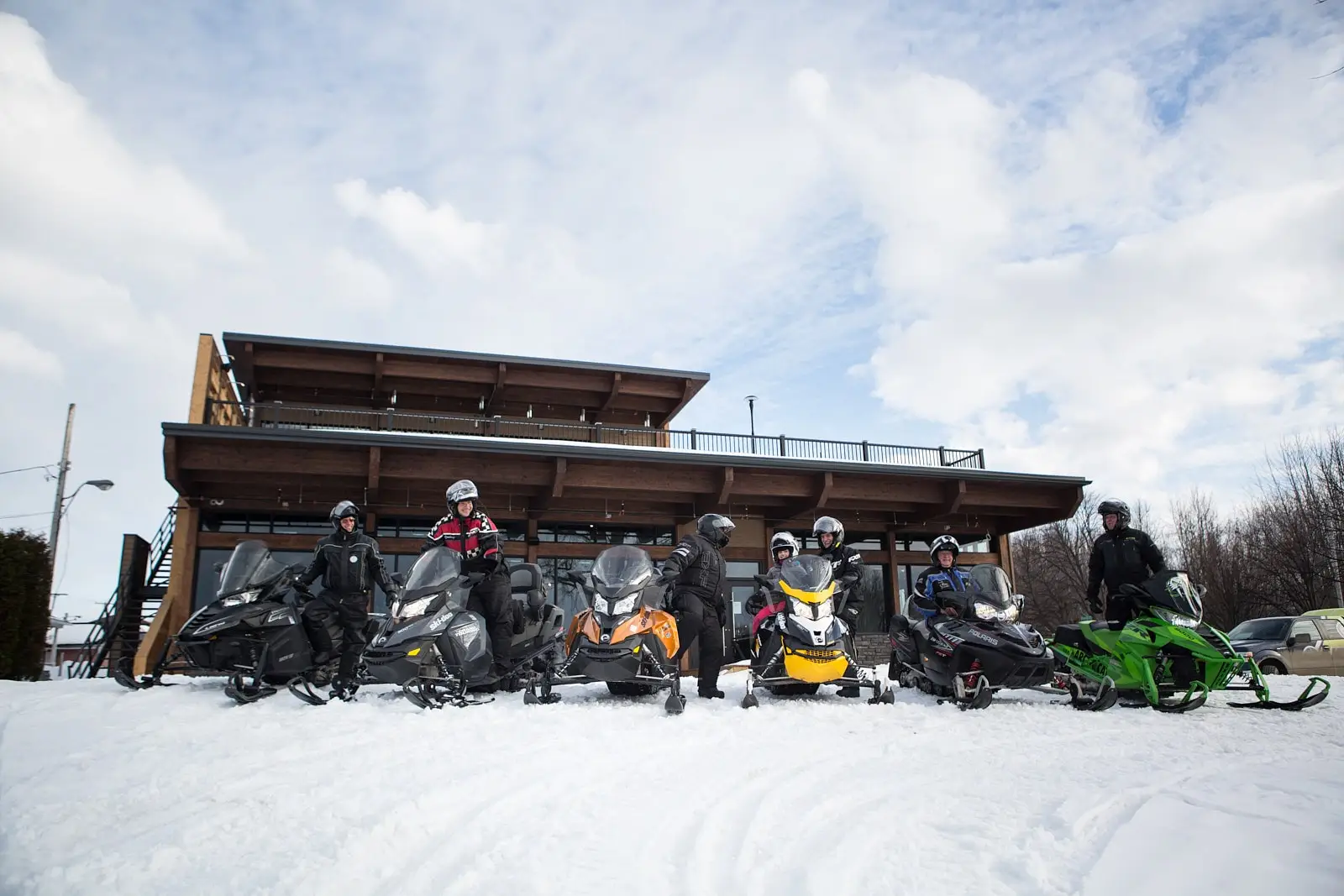 A group of snowmobilers posing with their colourful snowmobiles in front of the modern wooden chalet under a cloudy sky in Centre-du-Québec.