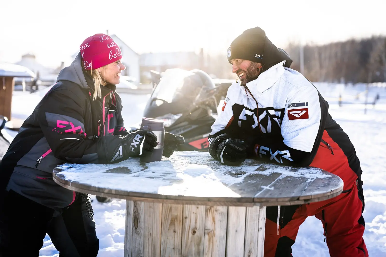 Two snowmobilers in FXR winter outfits sit around a snow-covered table, enjoying a hot drink with snowmobiles in the background.