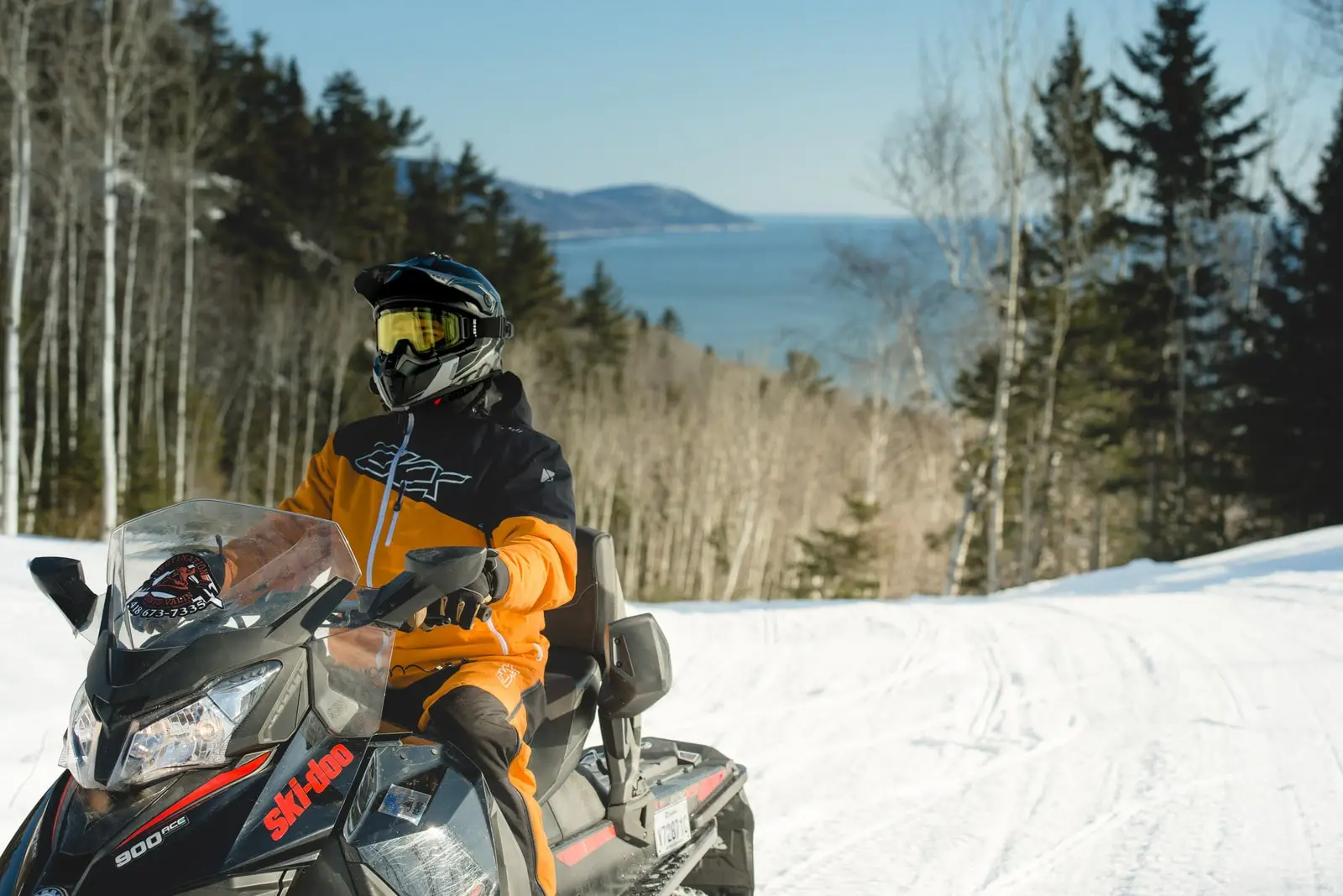 Motoneigiste en tenue orange sur une piste enneigée avec vue sur un paysage montagneux et maritime.