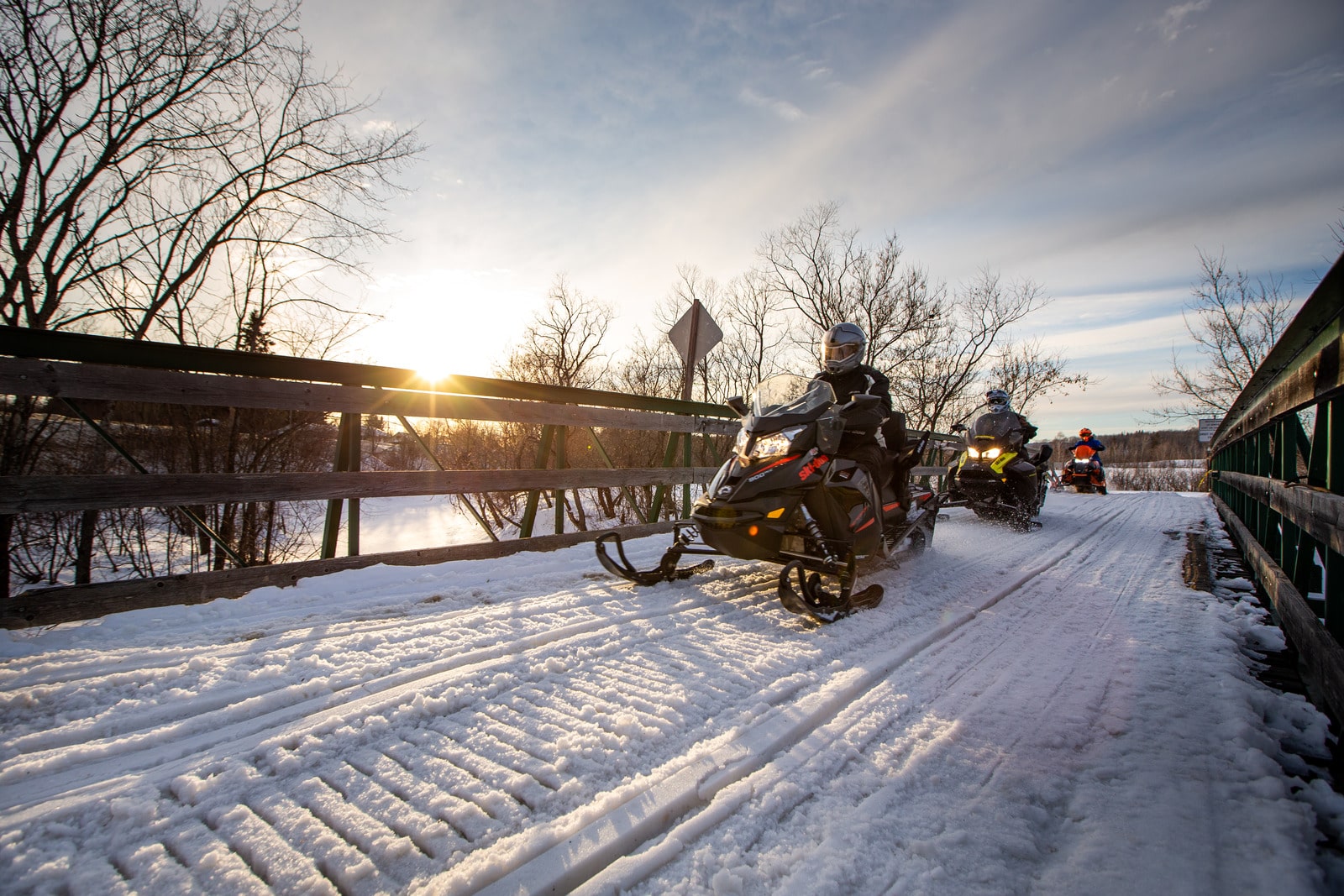 Groupe de motoneigistes traversant un pont enneigé au coucher du soleil, sur un sentier de motoneige au Québec.