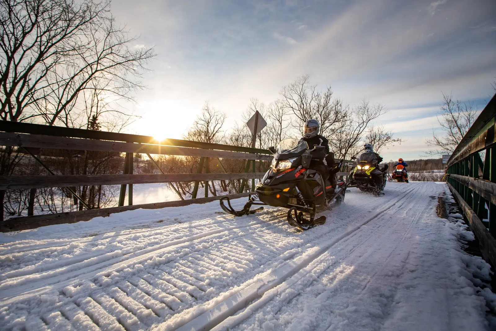 Snowmobilers crossing a snow-covered bridge at sunset on a snowmobile trail in Québec.