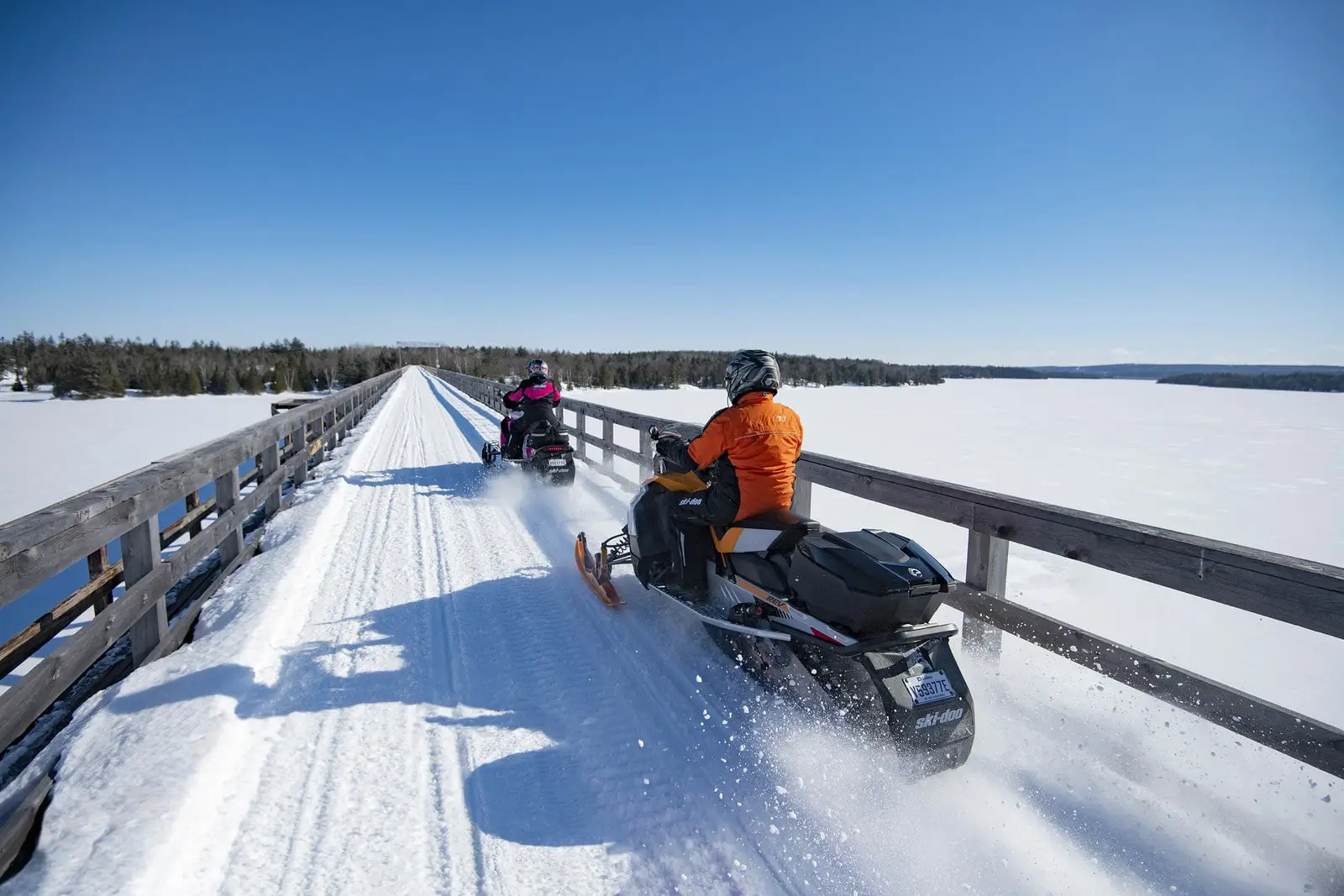 Motoneigistes traversant un pont enneigé au-dessus d’un lac gelé au Québec.