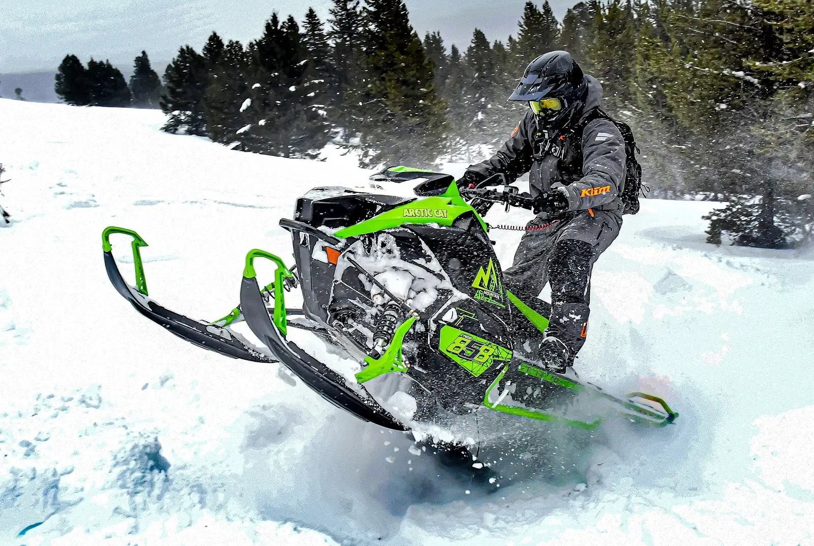 Rider maneuvering a green M 858 Alpha One Sno Pro Arctic Cat snowmobile through deep powder snow, kicking up a spray of snow with pine trees in the background.