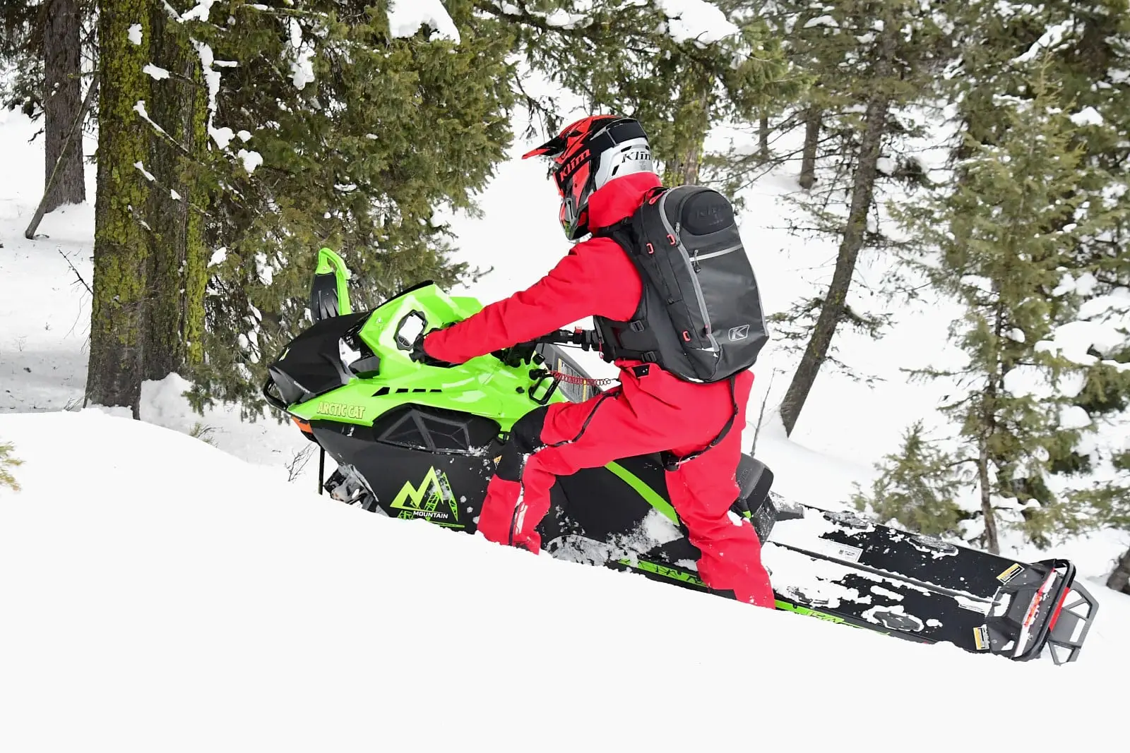 Rider in red gear navigating a green Arctic Cat snowmobile uphill through fresh snow, surrounded by snow-covered trees.