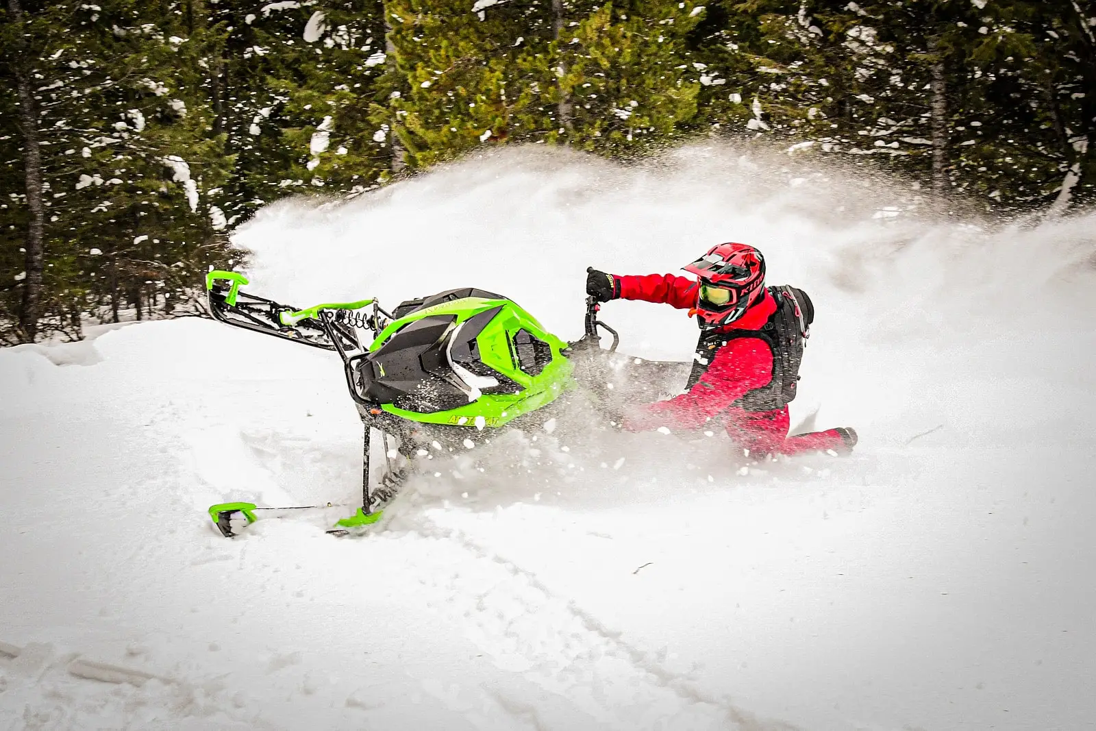 Rider performing a sharp turn on a green M 858 Alpha One Sno Pro snowmobile, creating an impressive snow spray against a forested backdrop.