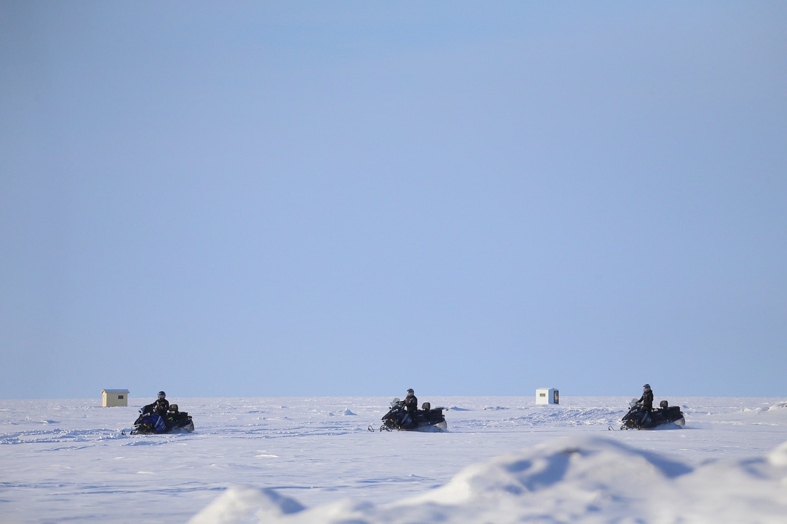 Motoneiges traversant un lac gelé bordé de cabanes, dans les sentiers de motoneige du Saguenay–Lac-Saint-Jean.