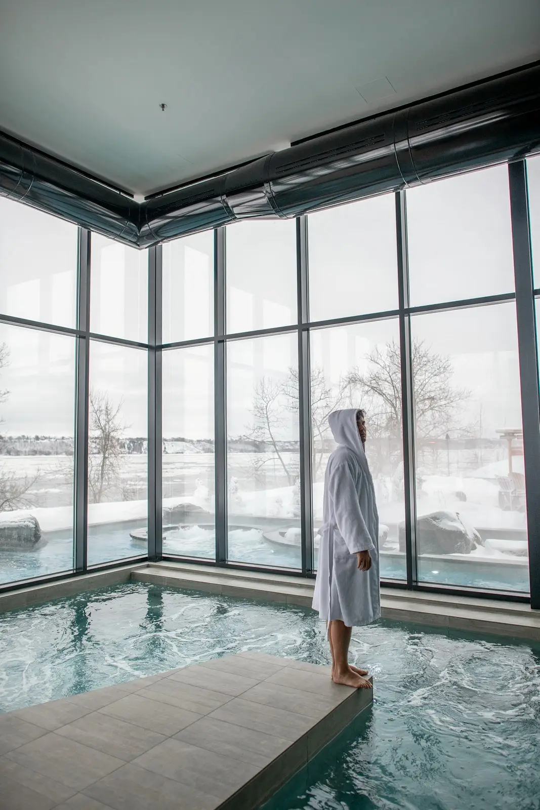 Une piscine intérieure paisible avec une personne en peignoir blanc regardant à travers de grandes fenêtres sur un paysage enneigé.