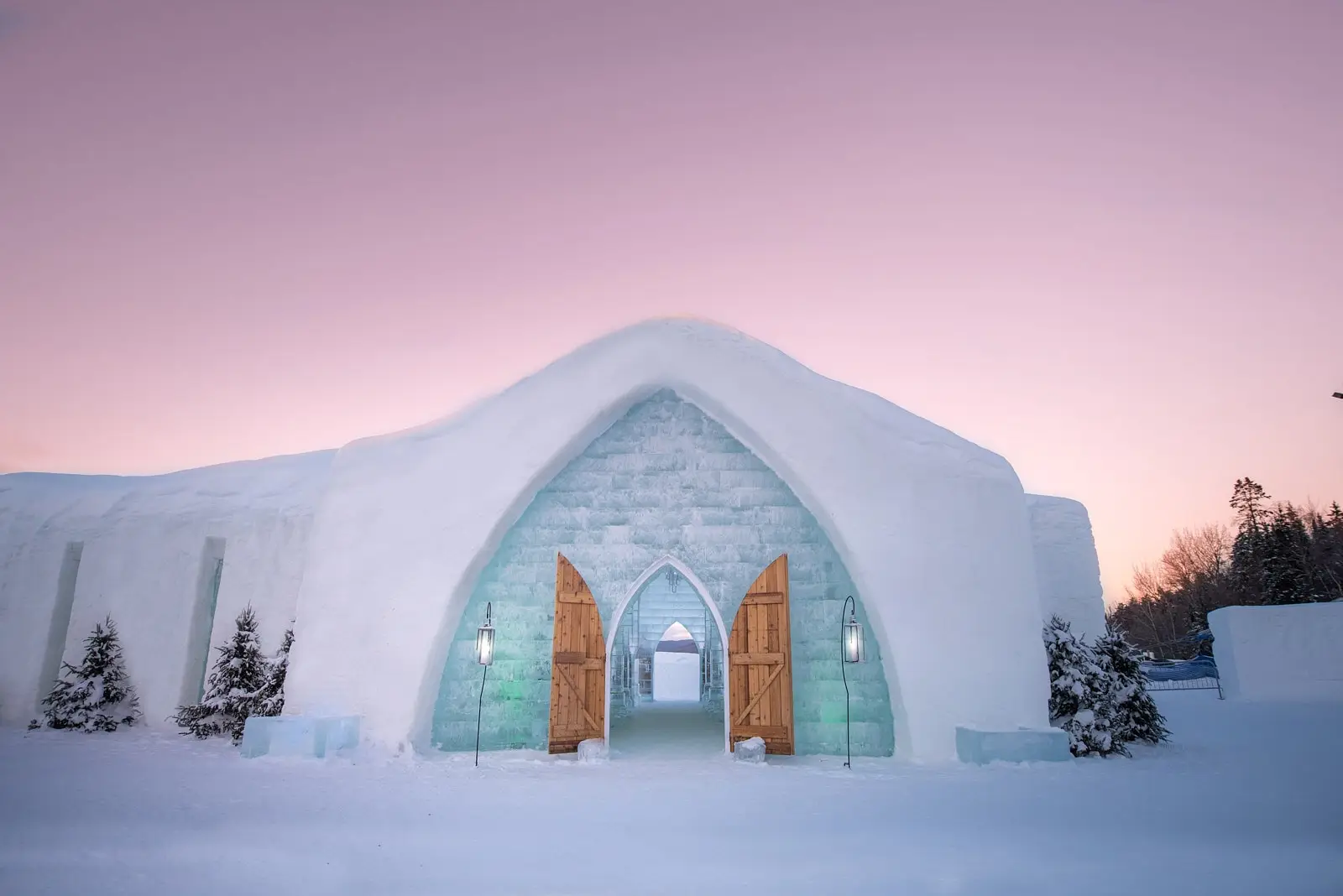 L’entrée de l’Hôtel de Glace, avec des portes en bois arquées et des murs de glace, sous un ciel dégradé rose et violet au coucher du soleil.