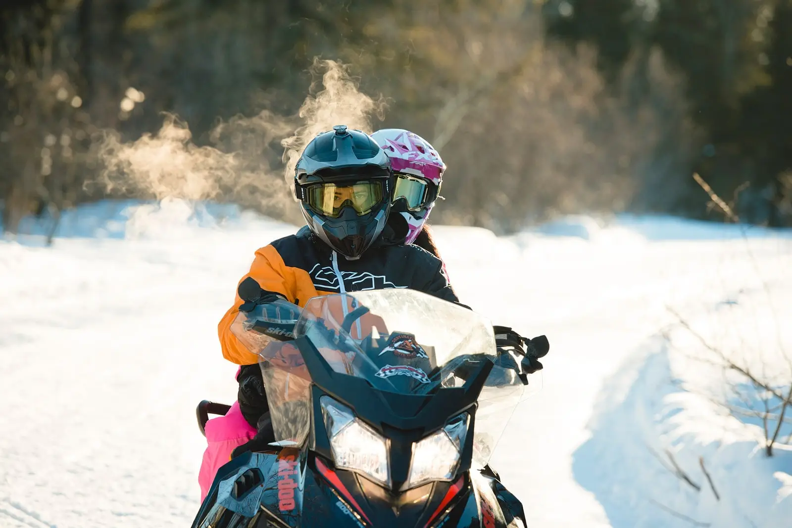 A snowmobile with two passengers riding along a snowy trail, steam rising from their helmets in the crisp winter air.