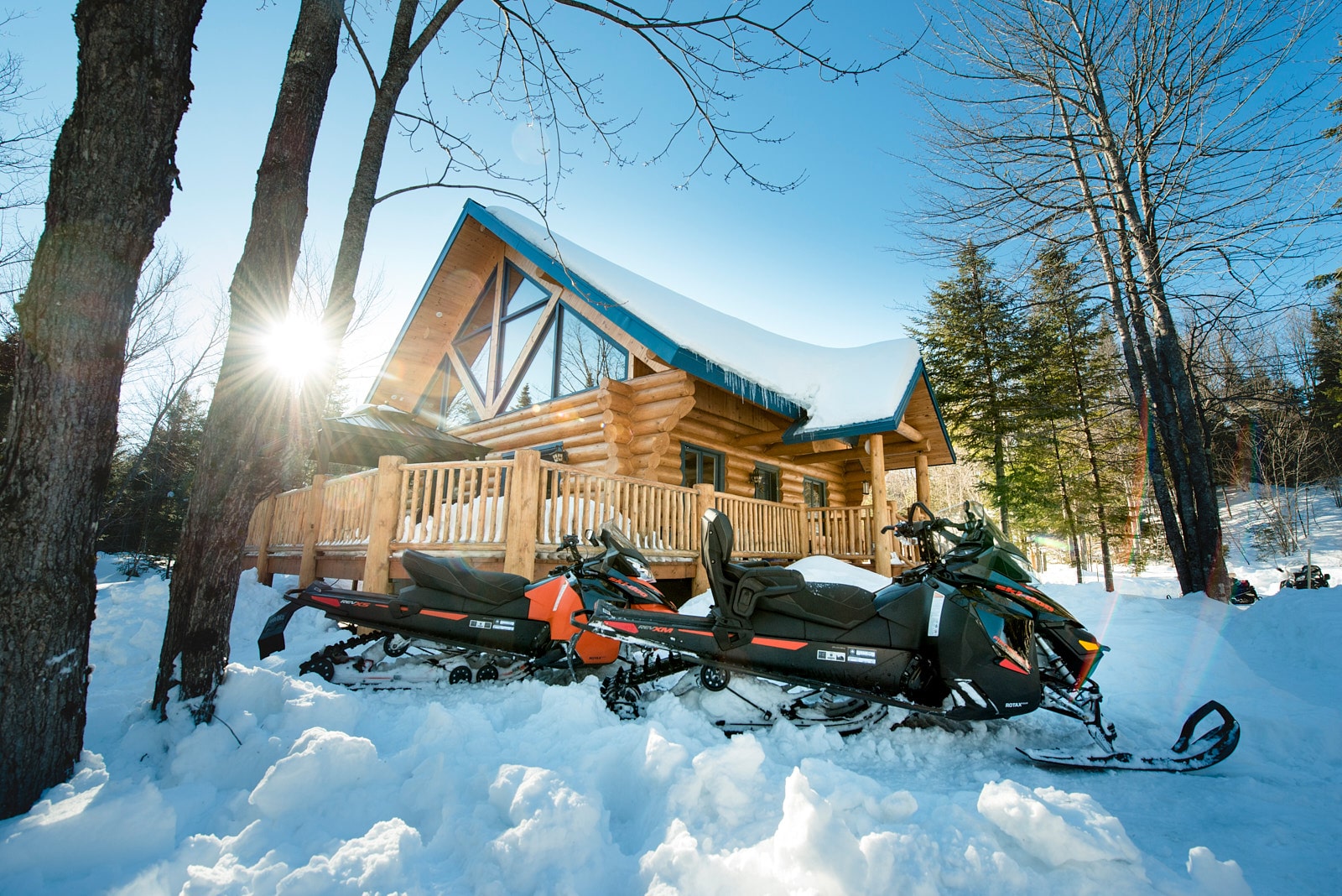 Un chalet en bois rond avec un toit couvert de neige, entouré d’arbres et de motoneiges stationnées, éclairé par une vive lumière hivernale.