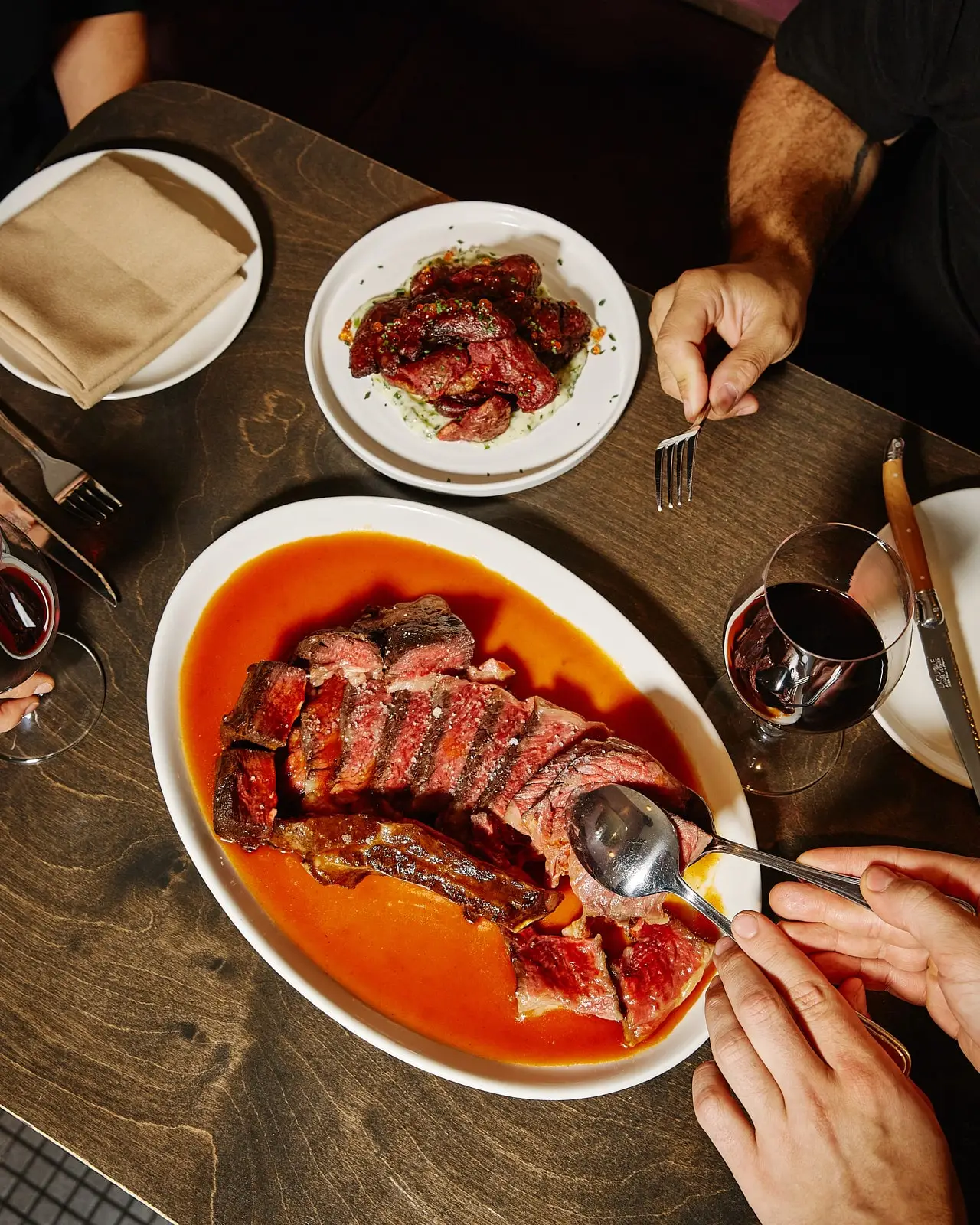 Une table partagée avec un grand plat de steak saignant tranché dans une sauce rouge, un accompagnement de viande rôtie et des verres de vin rouge.