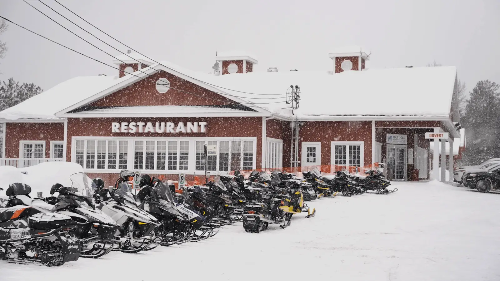  In front of a red wooden restaurant covered with snow in Lanaudière-Mauricie, a row of snowmobiles is parked while the snow falls slowly.