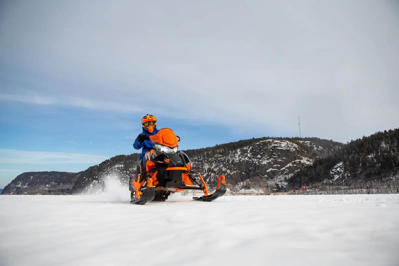 A snowmobile rider in a blue suit and orange helmet is driving an orange snowmobile across snow-covered terrain in Lanaudière-Mauricie, with wooded hills in the background.