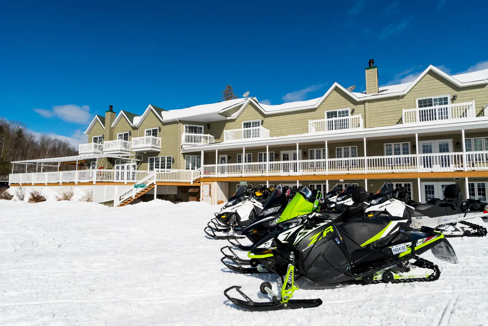 A row of snowmobiles parked in front of Pourvoirie du Lac Blanc, a large green two-storey building with balconies, under a clear blue sky.