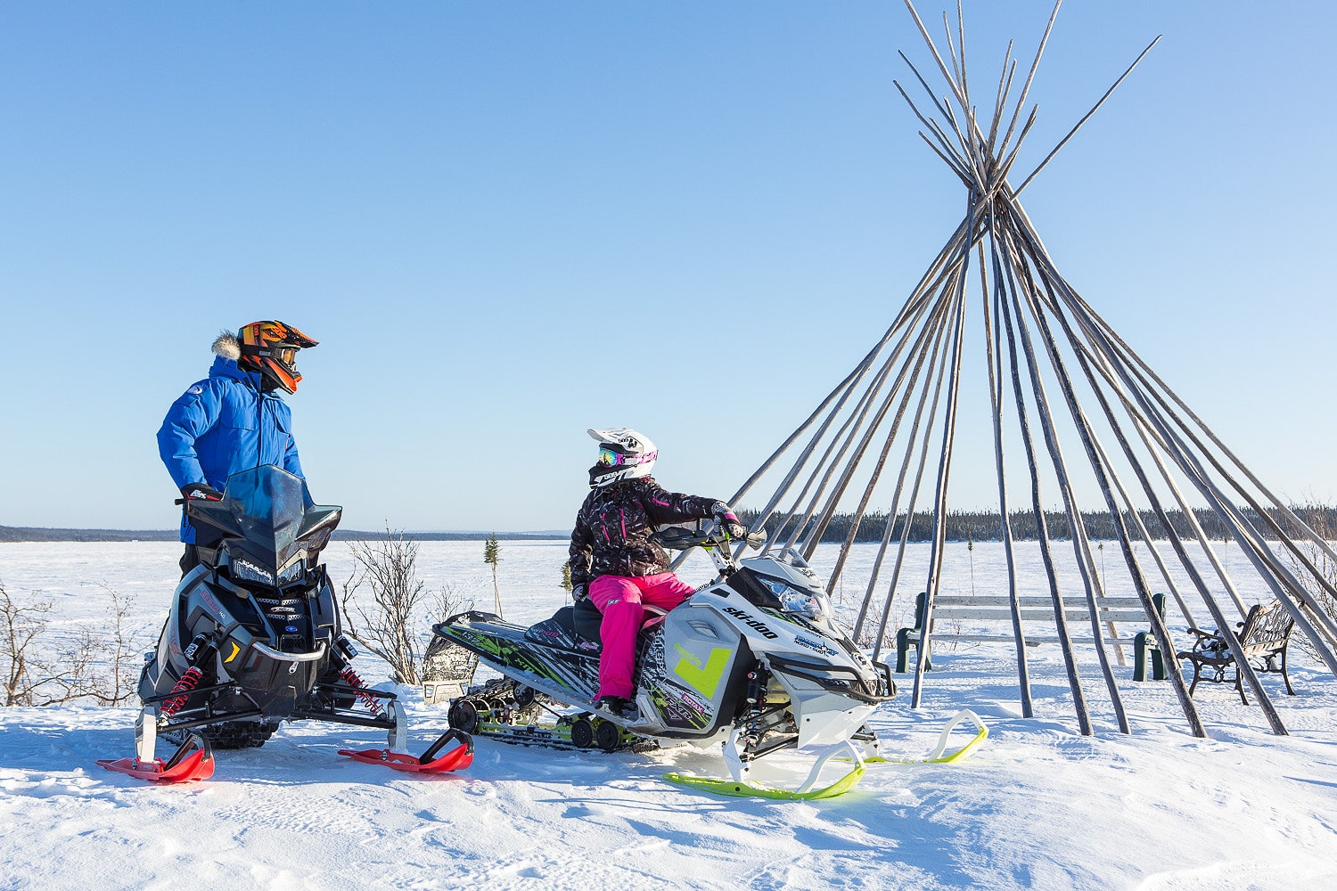 Deux motoneigistes à l’arrêt, l’un sur une Ski-Doo blanche et l’autre sur une motoneige noire, près d’une structure de tipi en bois sur un terrain enneigé avec un ciel bleu dégagé en arrière-plan.