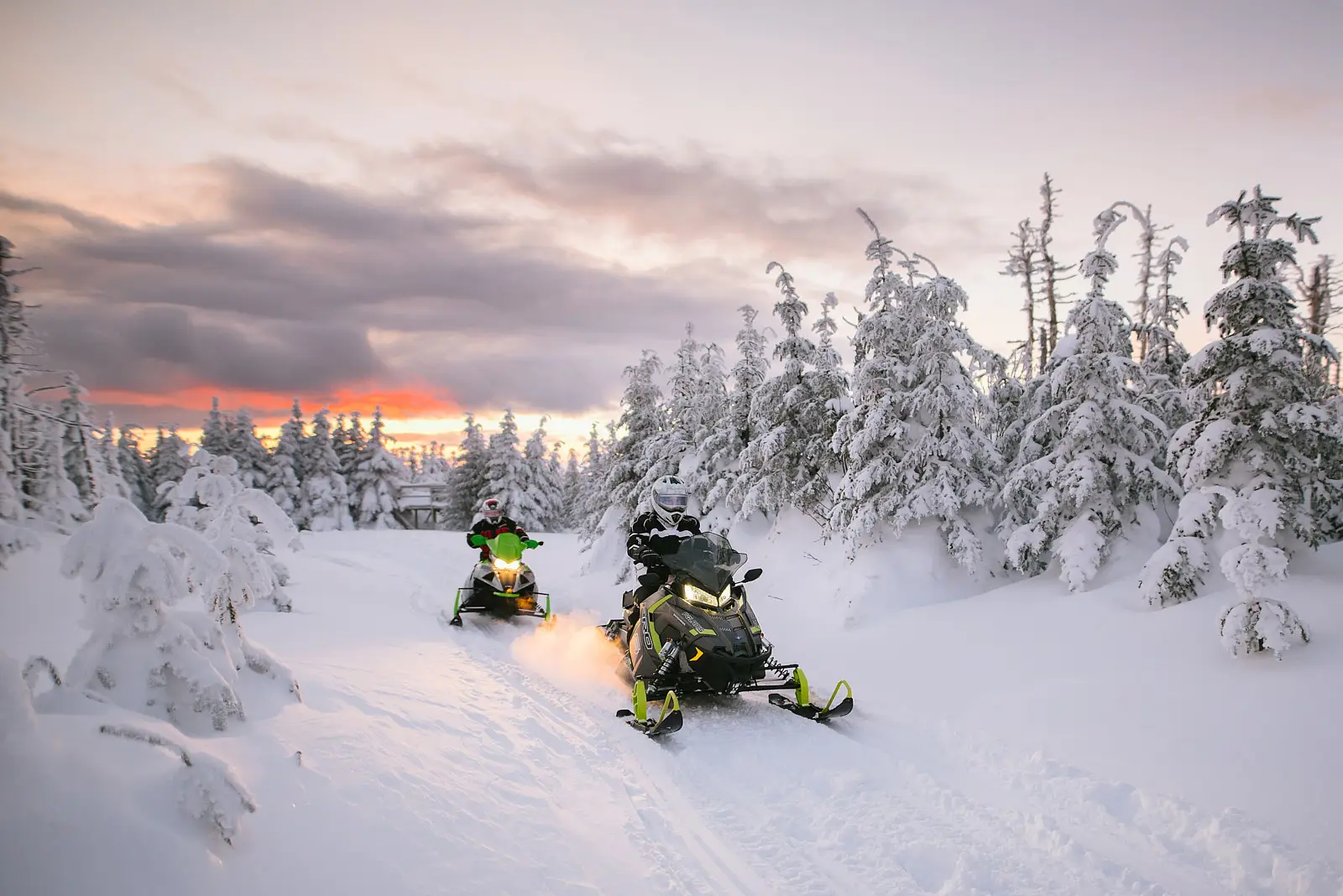 Two snowmobiles on a snowy trail surrounded by snow-covered trees, with a vibrant sunset sky in the background.